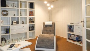 Una stanza con un lettino per massaggi, scaffali con libri e oggetti decorativi, e una scrivania bianca con un libro.