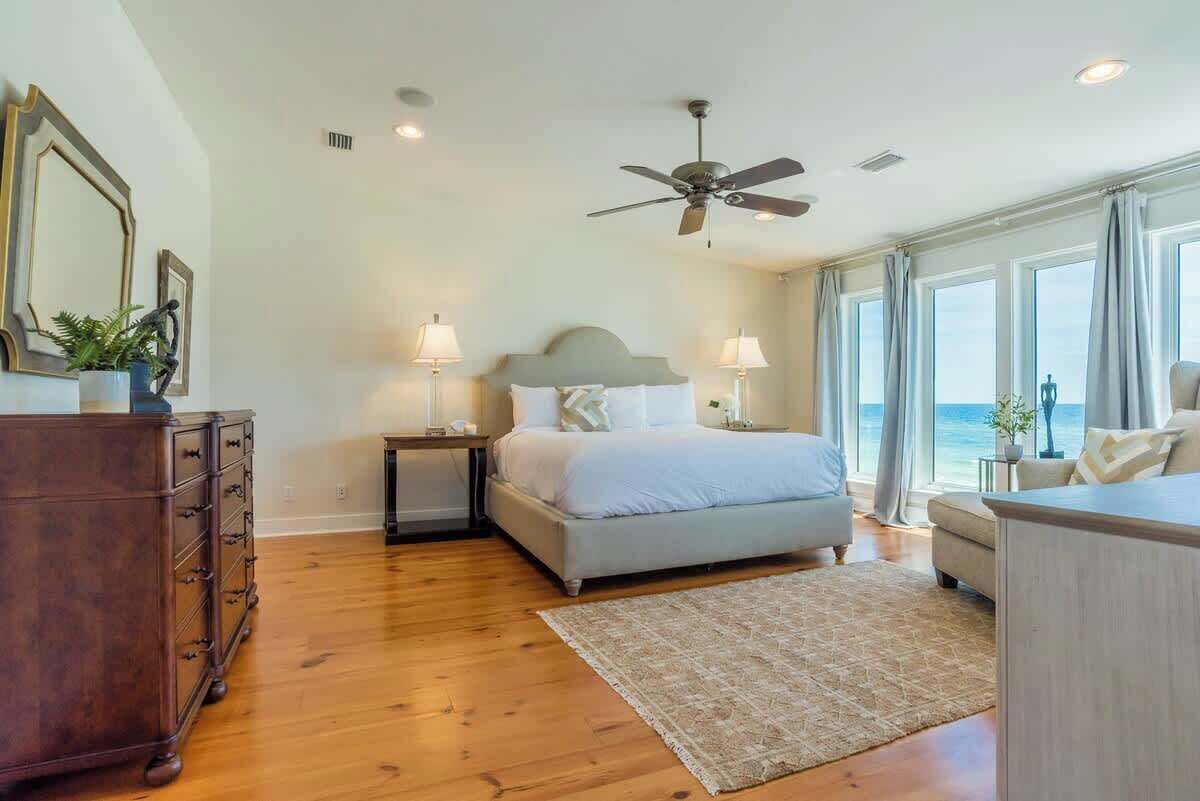 A bedroom with a bed, bedside tables, a dresser, a ceiling fan, and large windows with curtains.
