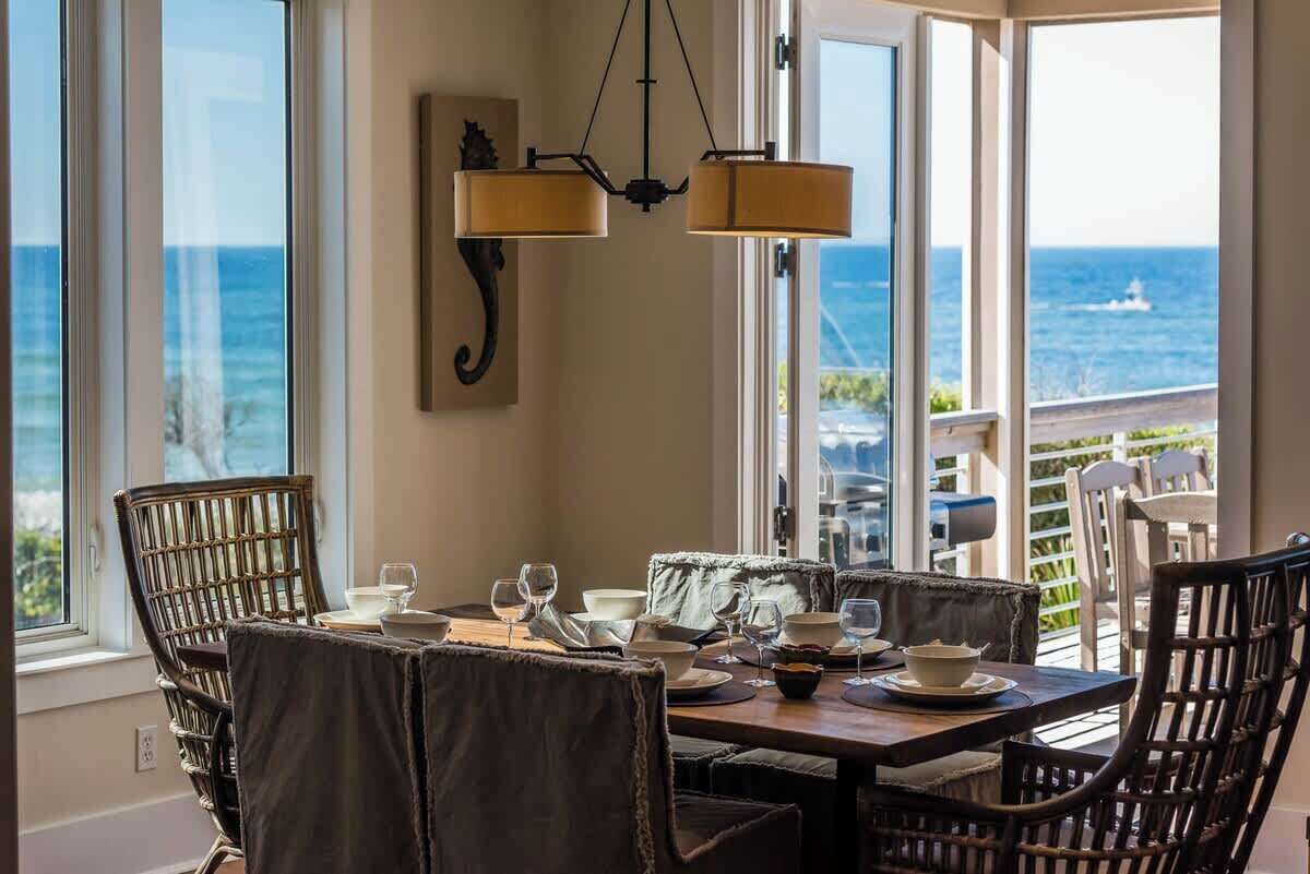 A dining area with a view of the ocean, featuring wicker chairs, a wooden table, and a hanging light fixture.