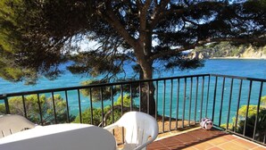 A balcony with white plastic chairs and a table, overlooking a coastal area with a tree and a view of the sea.