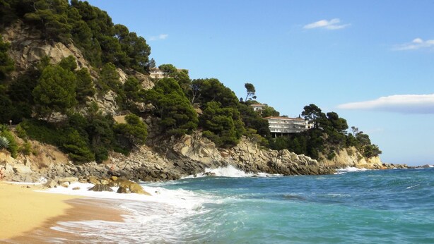 A coastal landscape with a beach, rocky cliffs, and a building with a red roof.