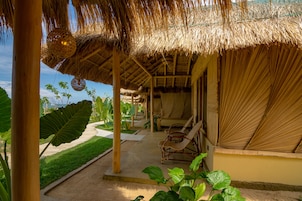 Un área de estar al aire libre con techo de paja, sillones de descanso, rodeada de plantas tropicales y una vista despejada del océano.