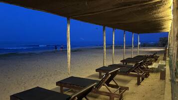 Un área de descanso frente a la playa con sillones y mesas de madera bajo una estructura cubierta, con vistas al océano.