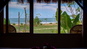 A room with a view of the beach through large windows, featuring a wicker chair and a small table with a vase. - Blue Waters Arugambay (Pottuvil)