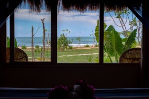 Habitación con vistas a la playa a través de amplios ventanales, que incluye una silla de mimbre y una mesita con un florero.