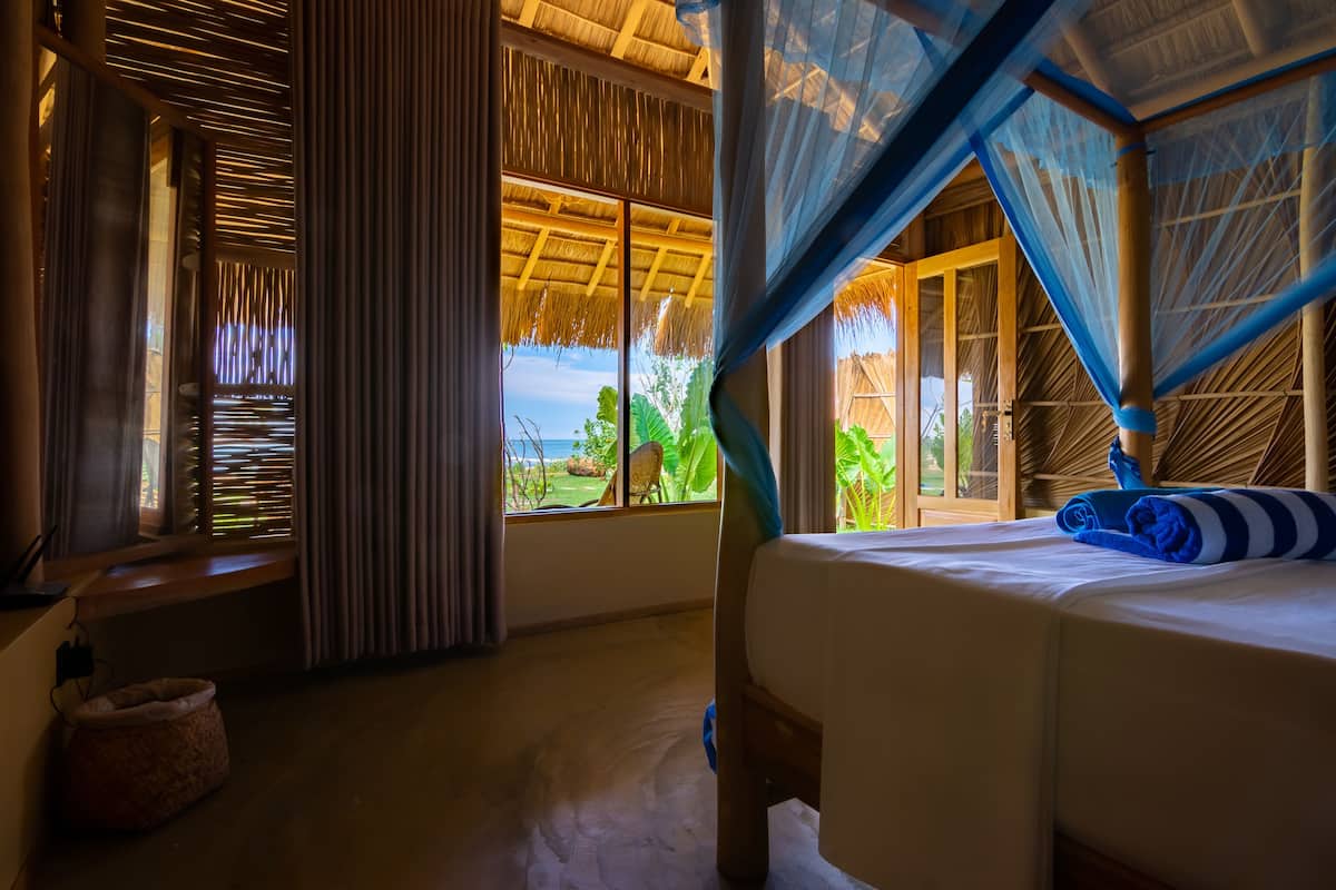 A room with a bed, a window with curtains, a thatched roof, and a view of the outdoors.