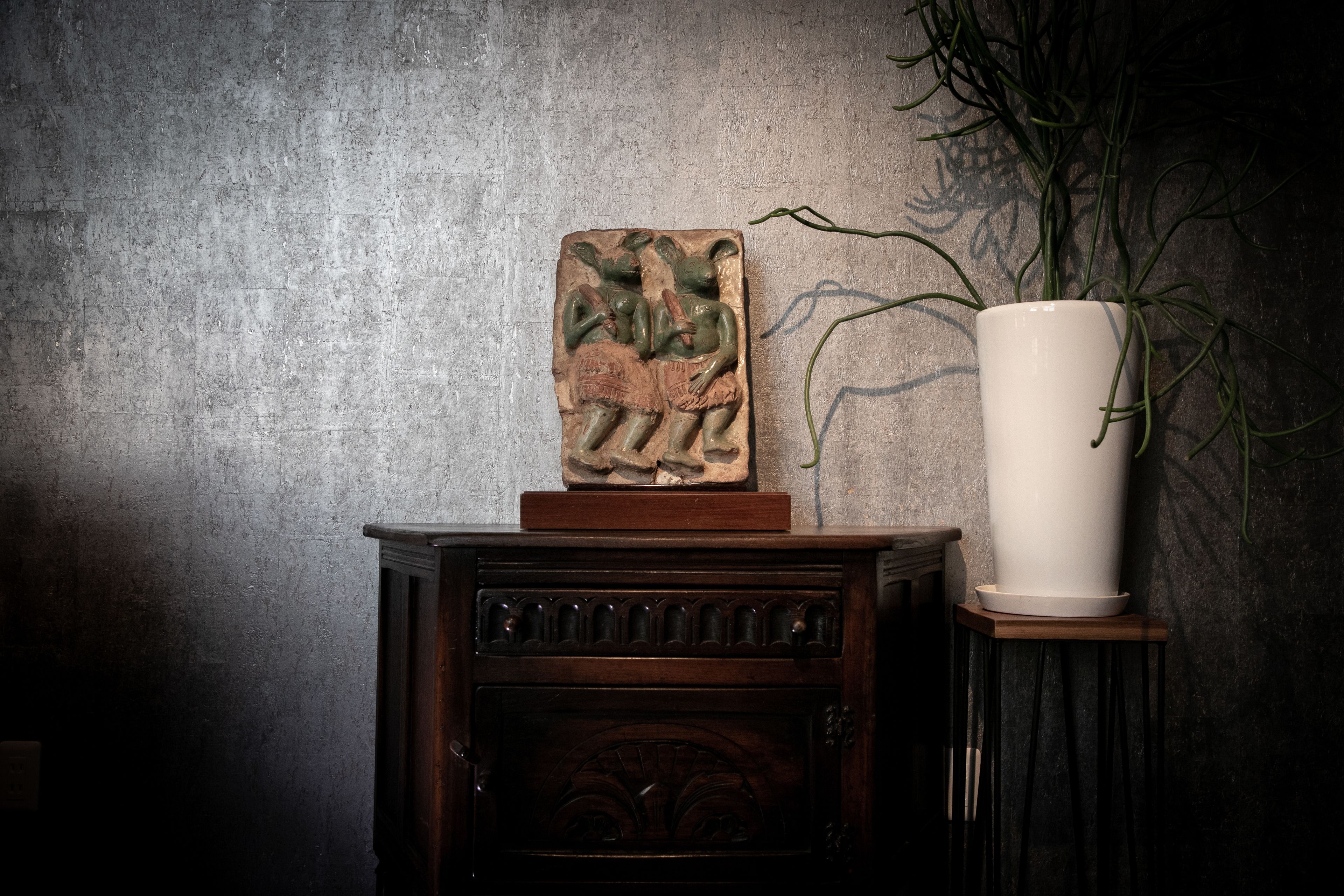Un meuble en bois rustique, orné d’une sculpture en relief sur le dessus, un vase blanc contenant une plante, posé sur une petite table à côté, et un mur gris texturé en arrière-plan.