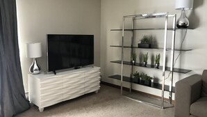 A modern living room with a flat-screen TV on a white console, a glass shelving unit with plants, and a grey sofa. - Fully Renovated 2 Bed, 1 Bath Apartment near 8th Street (Saskatoon)