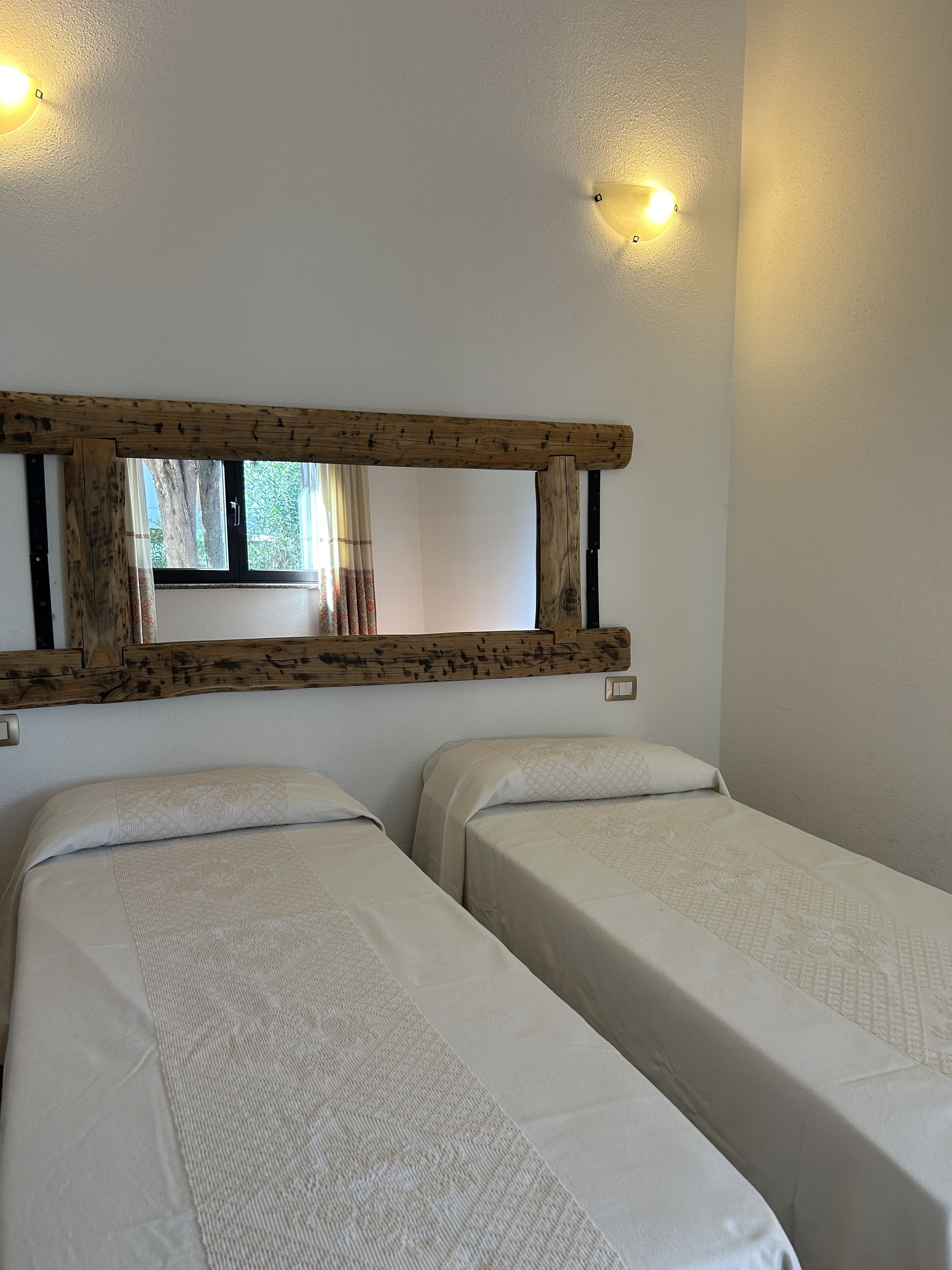 Een kamer met twee bedden, een houten hoofdeinde, een raam met gordijnen en wandlampen.