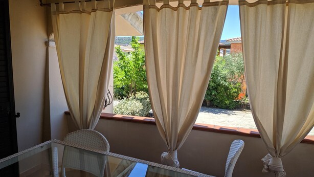 Un balcón con una mesa de vidrio y dos sillas de mimbre, con vistas a un jardín con árboles y un edificio al fondo.