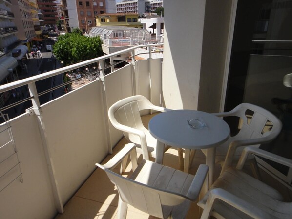 A balcony with white plastic chairs and a small table, overlooking a street with buildings and a green tree.