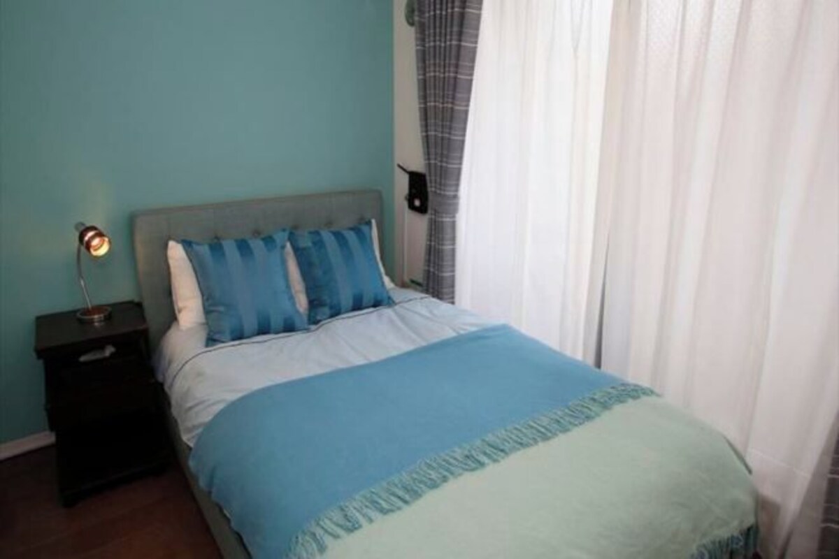 A bedroom with a bed, a nightstand, a wall-mounted lamp, and a window with curtains.