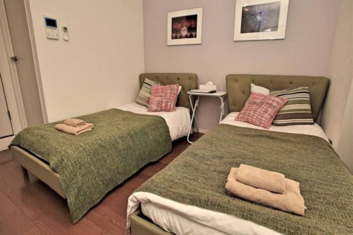 A twin room with two beds, each with a green blanket and a folded towel. There is a small bedside table with a lamp and a picture frame on the wall above the beds.