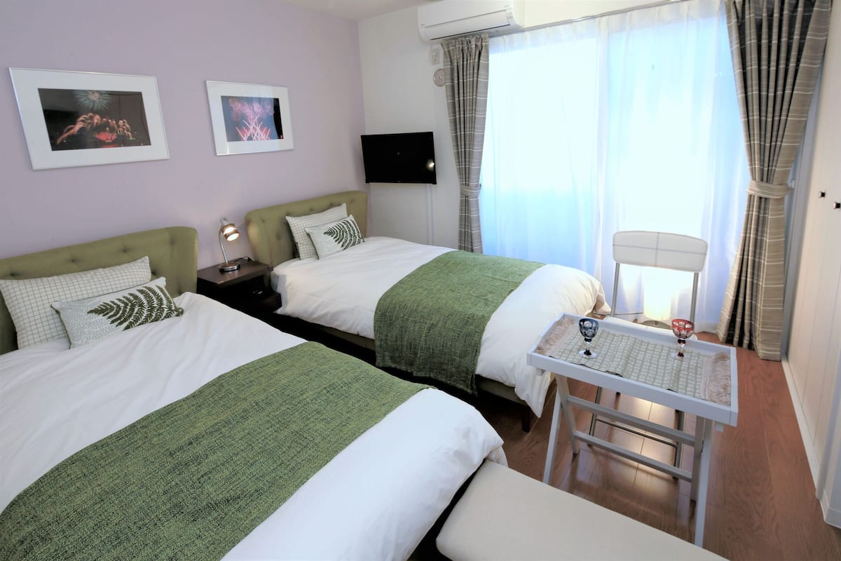 A hotel room with two beds, a flat-screen TV, a small table with decorative items, and a window with curtains.