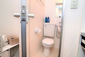 Une petite salle de bain compacte, équipée de toilettes, d’un lavabo et d’un miroir. On trouve des articles de toilette sur le lavabo et un panier sur le sol.