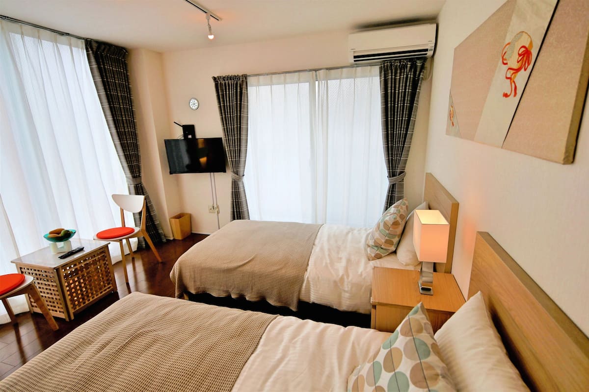 A hotel room with two beds, a TV, a bedside table with a lamp, and a window with curtains.