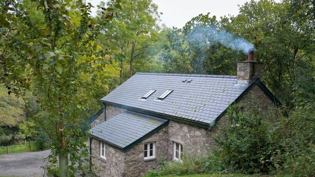 A stone house with a sloped roof, a chimney emitting smoke, and a small attached structure with a slanted roof.