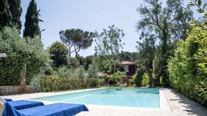 Une piscine avec des chaises longues, entourée de verdure et avec un bâtiment en arrière-plan.