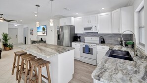 A modern kitchen with white cabinets, a marble countertop, and stainless steel appliances. There are wooden stools at the island and a ceiling fan. - The Beach House | Southern Exposure | Walk or Bike to Vanderbilt Beach (Naples)