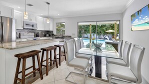 A modern kitchen with white cabinets, a stainless steel refrigerator, and a glass dining table with white chairs. - The Beach House | Southern Exposure | Walk or Bike to Vanderbilt Beach (Naples)