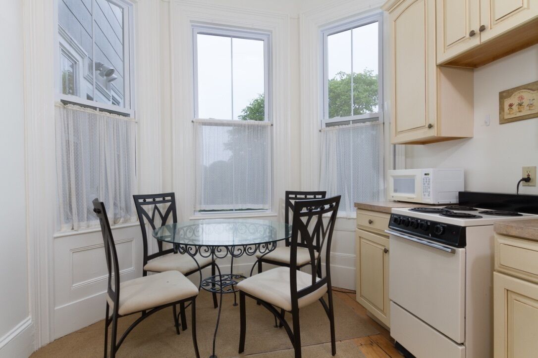 A kitchen with a round glass table, four chairs, a stove, a microwave, and a window with a view of trees.