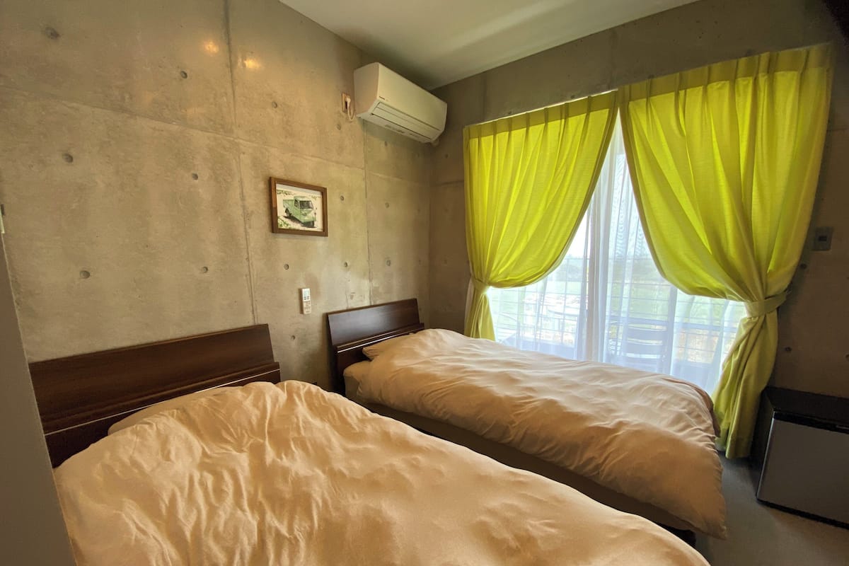 A hotel room with two beds, a wall-mounted air conditioner, a small picture, and a window with yellow curtains.