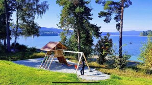 A wooden swing set with a metal frame and a ladder, situated on a gravel area surrounded by grass and trees, overlooking a lake with a clear sky. - HOUSE ON THE LAKE WITH DIRECT ACCESS TO THE LAKE (Frymburk nad Vltavou)