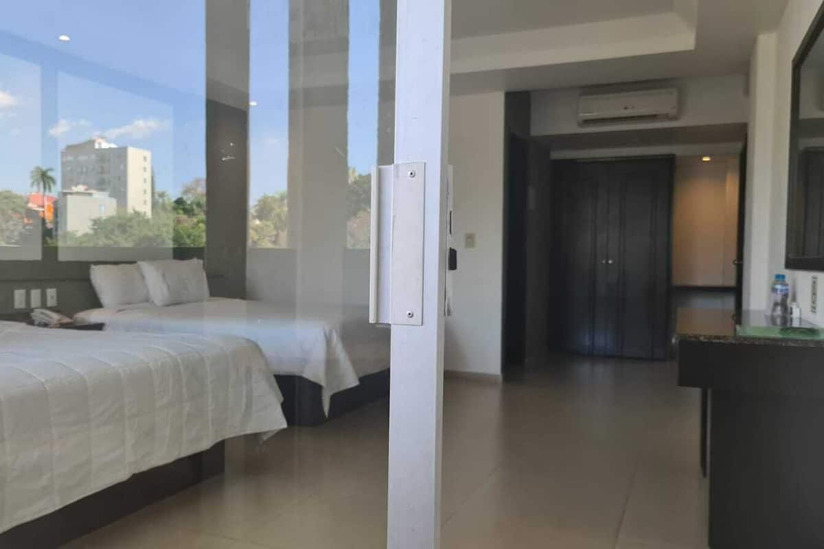 A hotel room with two beds, a glass partition, a view of the city, and a dark-toned door.