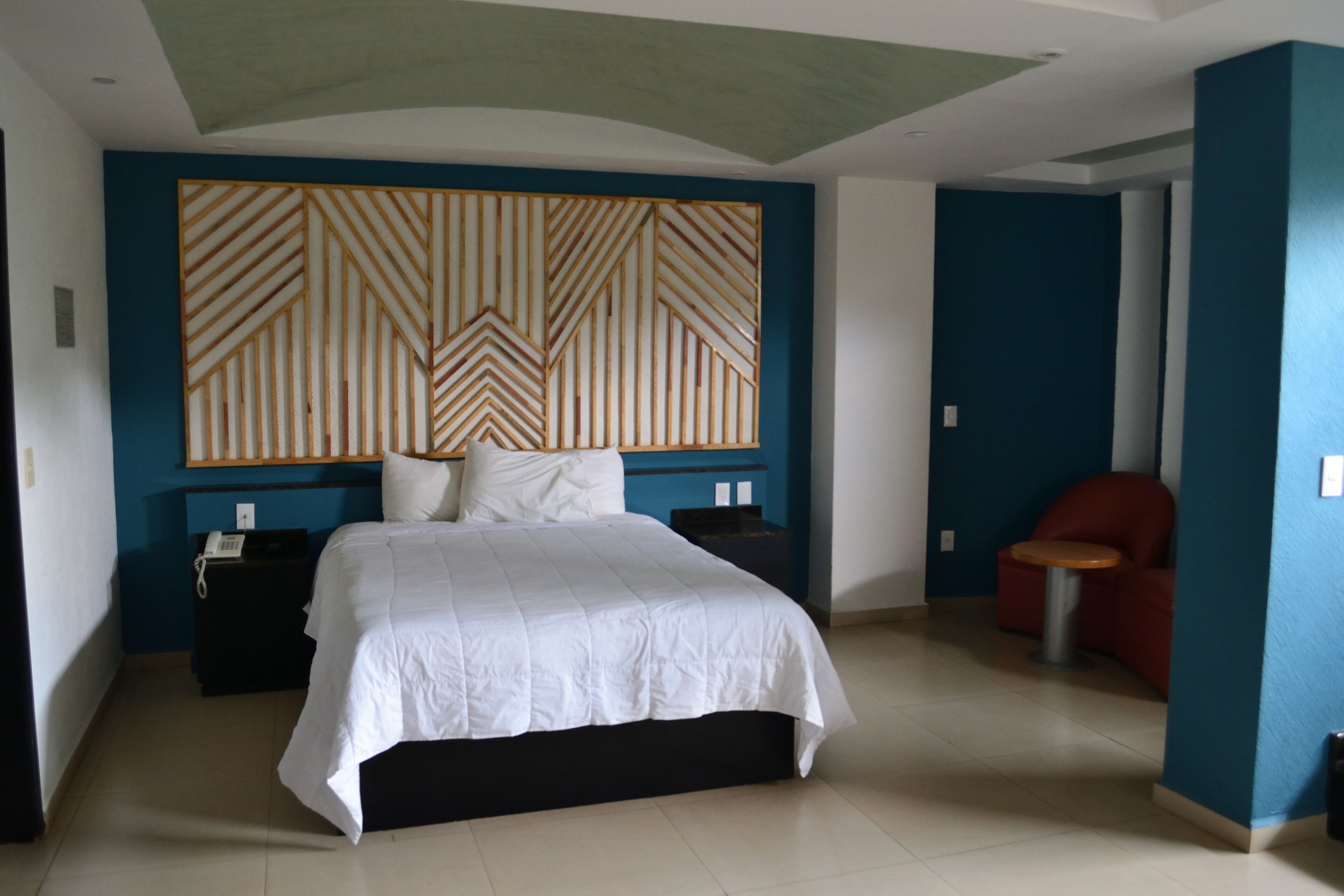 Habitación de hotel con una cama grande, cabecero de madera, una mesita y una silla roja.