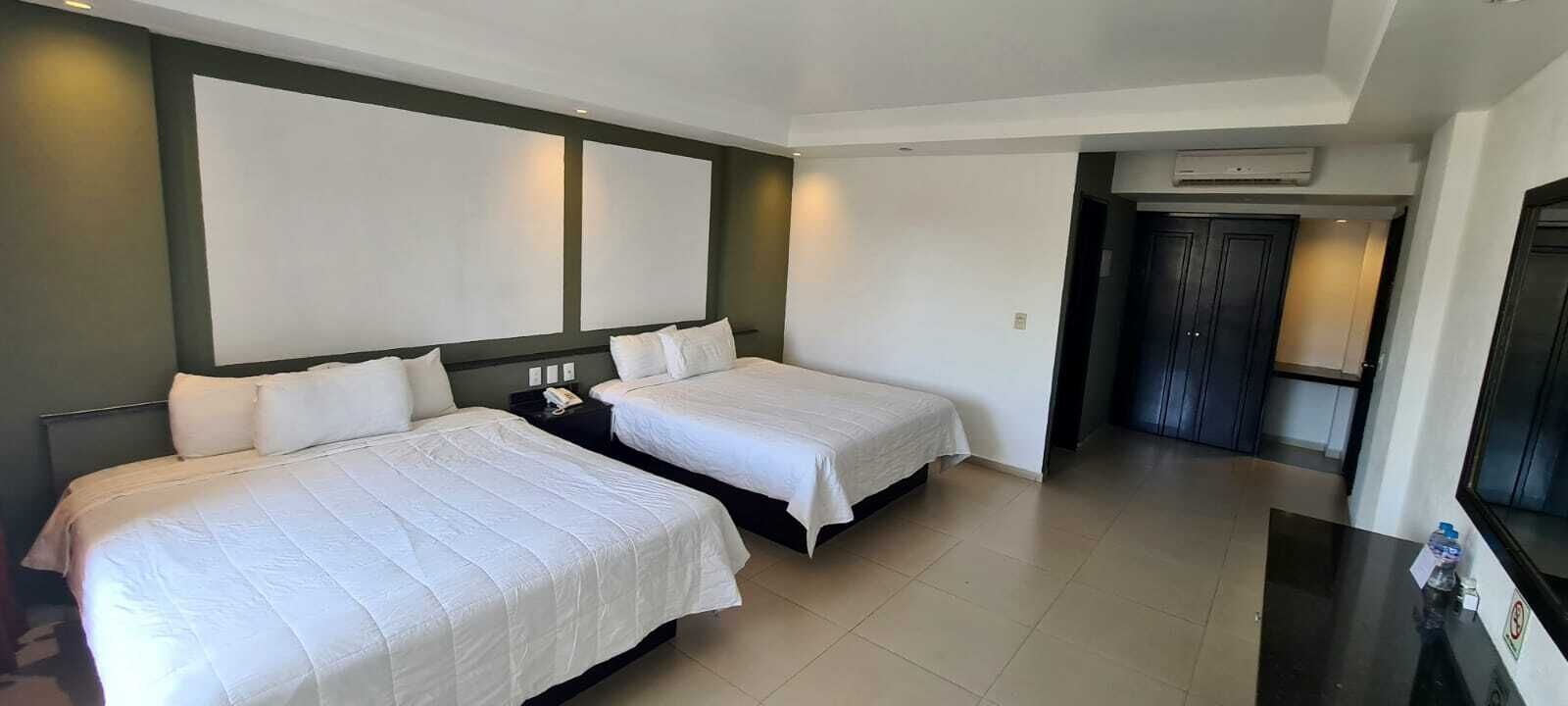 A hotel room with two beds, a dark wooden headboard, a small bedside table with a phone, and a door leading to a hallway.