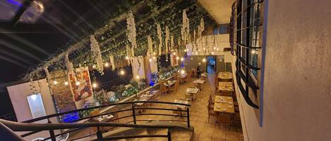 An indoor restaurant with a staircase leading to a dining area, decorated with hanging plants and string lights.