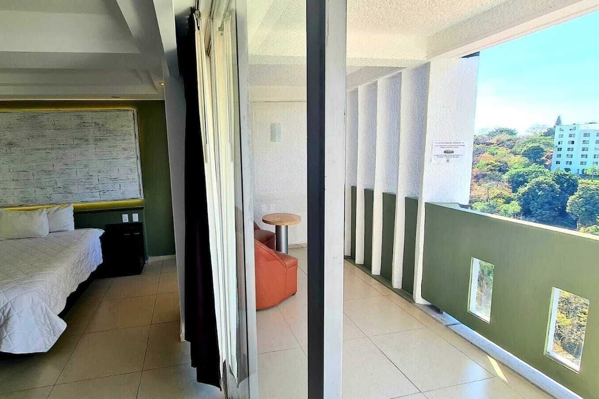A hotel room with a bed, a small table, and a balcony with a view of buildings and trees.