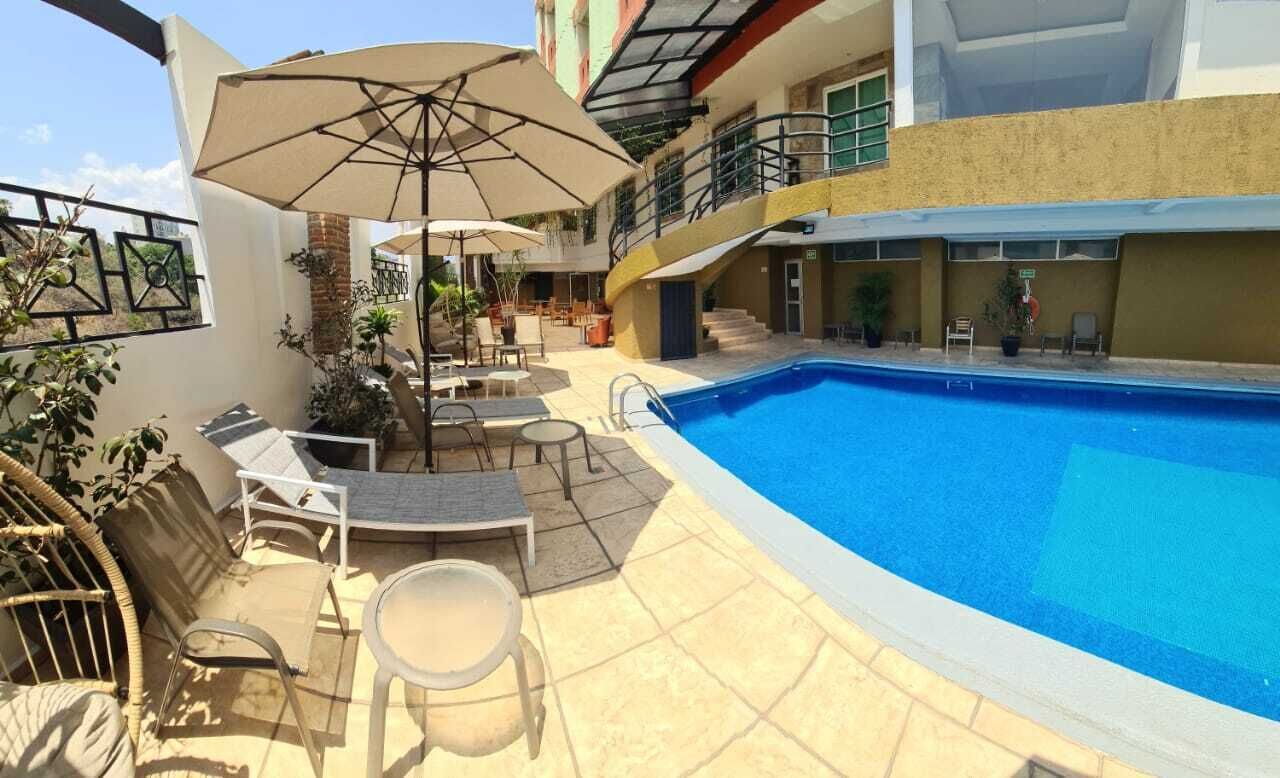 Une piscine entourée de chaises longues et de parasols, elle-même bordée d’un immeuble à plusieurs étages doté de balcons.