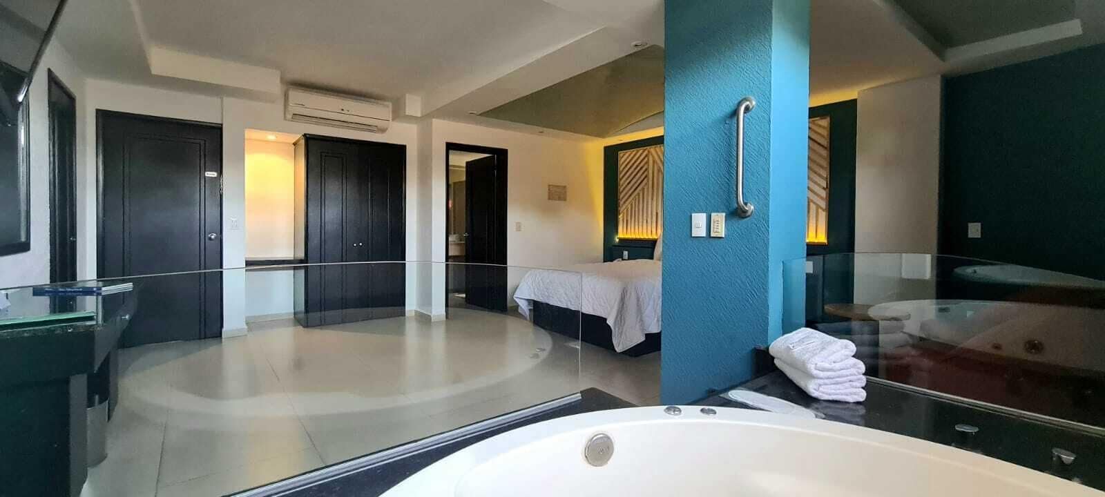 Una habitación de hotel moderna con una bañera grande, un dormitorio independiente con cama, y un baño con un espejo grande y un perchero para toallas.
