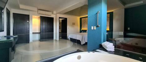 Una habitación de hotel moderna con una bañera grande, un dormitorio independiente con cama, y un baño con un espejo grande y un perchero para toallas.