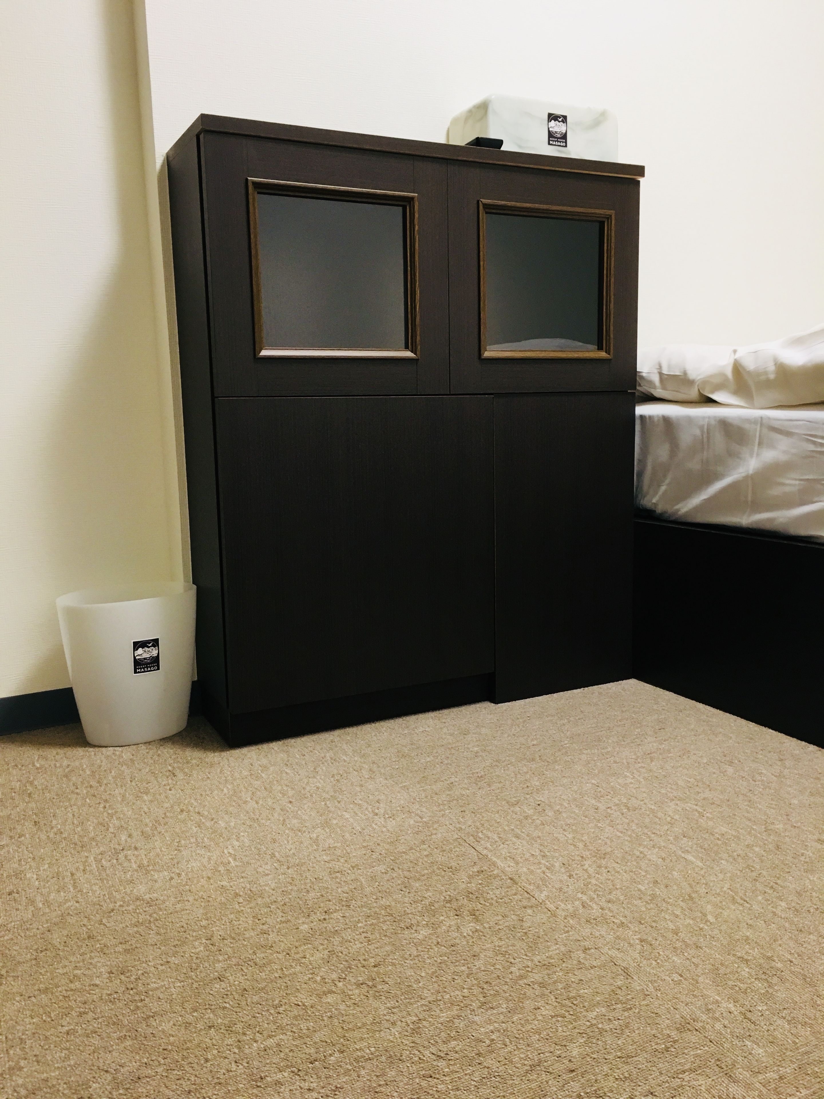 A dark wooden cabinet with two glass doors, a white trash bin, and a bed with white bedding.