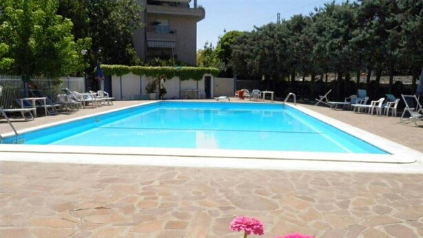 Uma piscina com um deck de pedra, rodeada por espreguiçadeiras e um edifício ao fundo.