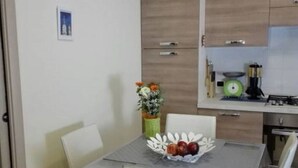 Uma sala de jantar com uma mesa posta para quatro pessoas, rodeada de cadeiras. Há um vaso com flores sobre a mesa, e uma cesta de frutas com maçãs. O espaço possui armários de madeira e uma área de cozinha com fogão e utensílios.