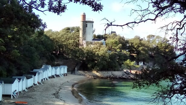Zona costera con un faro, cabañas de madera en la playa y una vista despejada del mar.