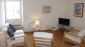 Una habitación minimalista con techo de madera, paredes blancas, un ventanal grande, una cama, un televisor y una mesita de noche con lámpara.