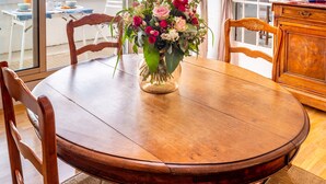 Une table à manger en bois, ornée d’un vase de fleurs, une chaise en bois et une vue sur la plage, visible à travers une porte vitrée.