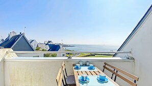 Un balcon avec une table dressée pour deux personnes, offrant une vue sur la côte, avec des bâtiments et la mer en toile de fond.