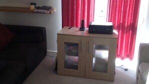 A small wooden cabinet with glass doors, a black electronic device on top, and a red curtain in the background. - King Size Bed in Large Room Kew Riverside Apartment  (Richmond)