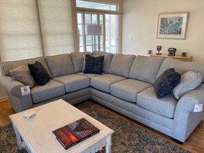 A grey sectional sofa with black cushions, a white coffee table with a book, and a framed picture on the wall.