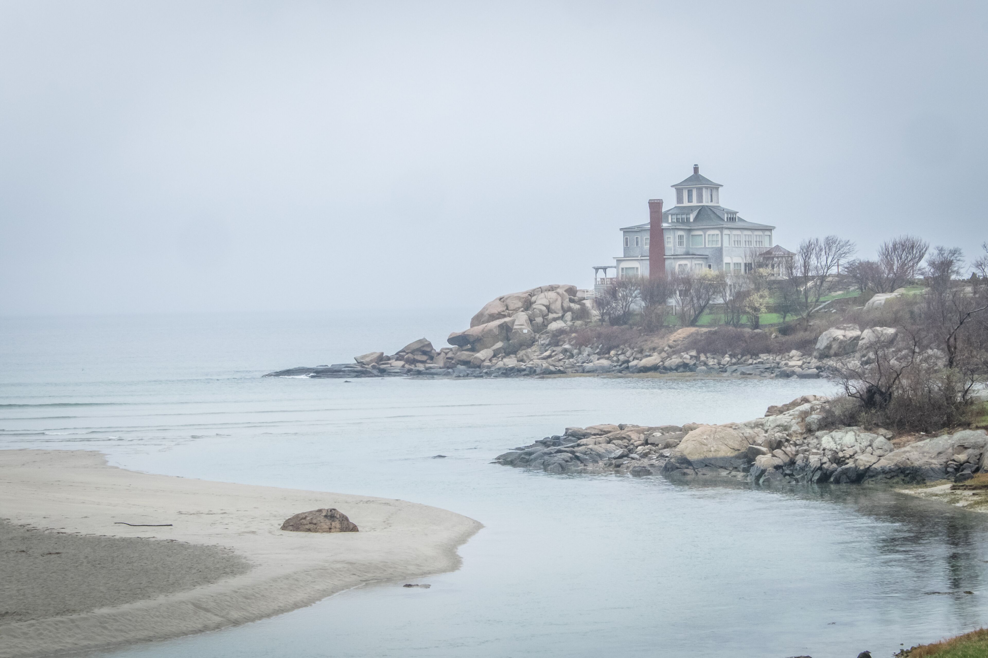Eine Küstenansicht mit einem großen weißen Gebäude auf einem Felsen, einem Sandstrand und ruhigem Wasser.