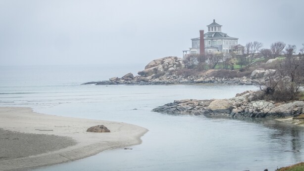 Paisatge costaner amb un edifici gran i blanc sobre un promontori rocos, una platja de sorra i aigua tranquil·la.