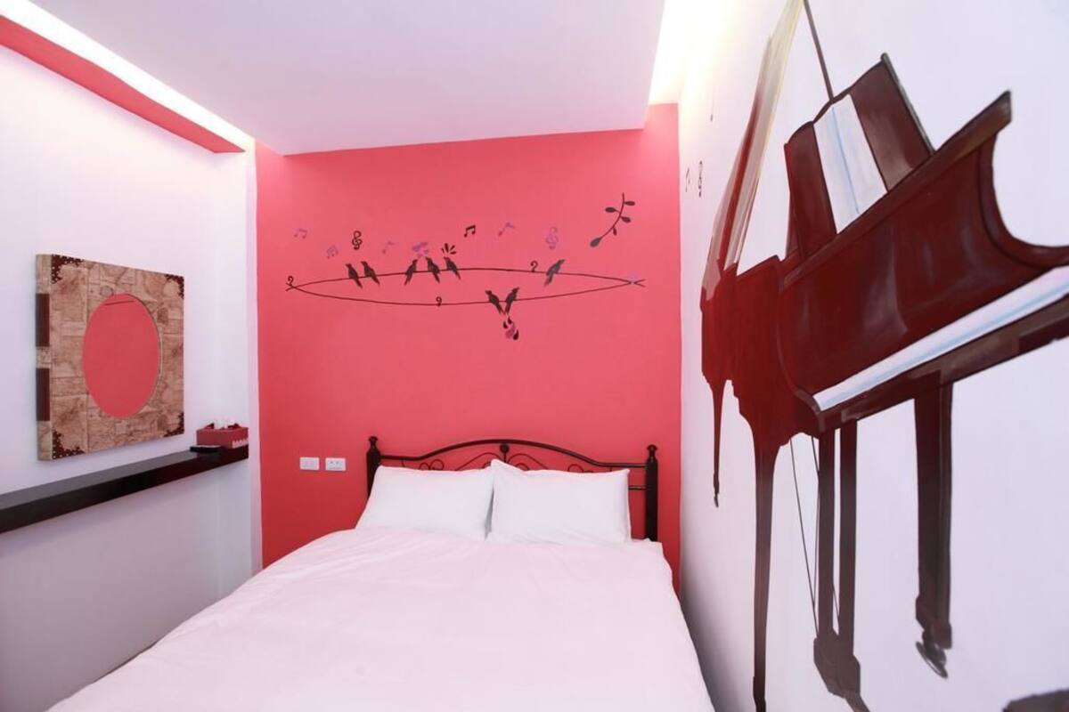 A bedroom with a red wall, a bed with white bedding, a black headboard, and a wall-mounted piano.