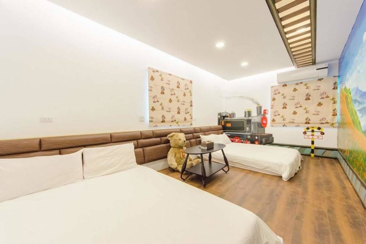 A hotel room with two beds, a small table, a teddy bear, and a mural on the wall.