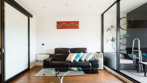 A modern living room with a glass coffee table, a black leather sofa with cushions, a red abstract painting on the wall, and a view of a balcony with a bicycle. - Cleo, 1BDR St Kilda Apartment (St Kilda)
