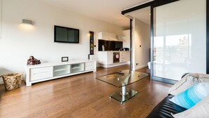 A modern living room with a glass coffee table, a white entertainment unit, and a kitchen in the background. - Cleo, 1BDR St Kilda Apartment (St Kilda)